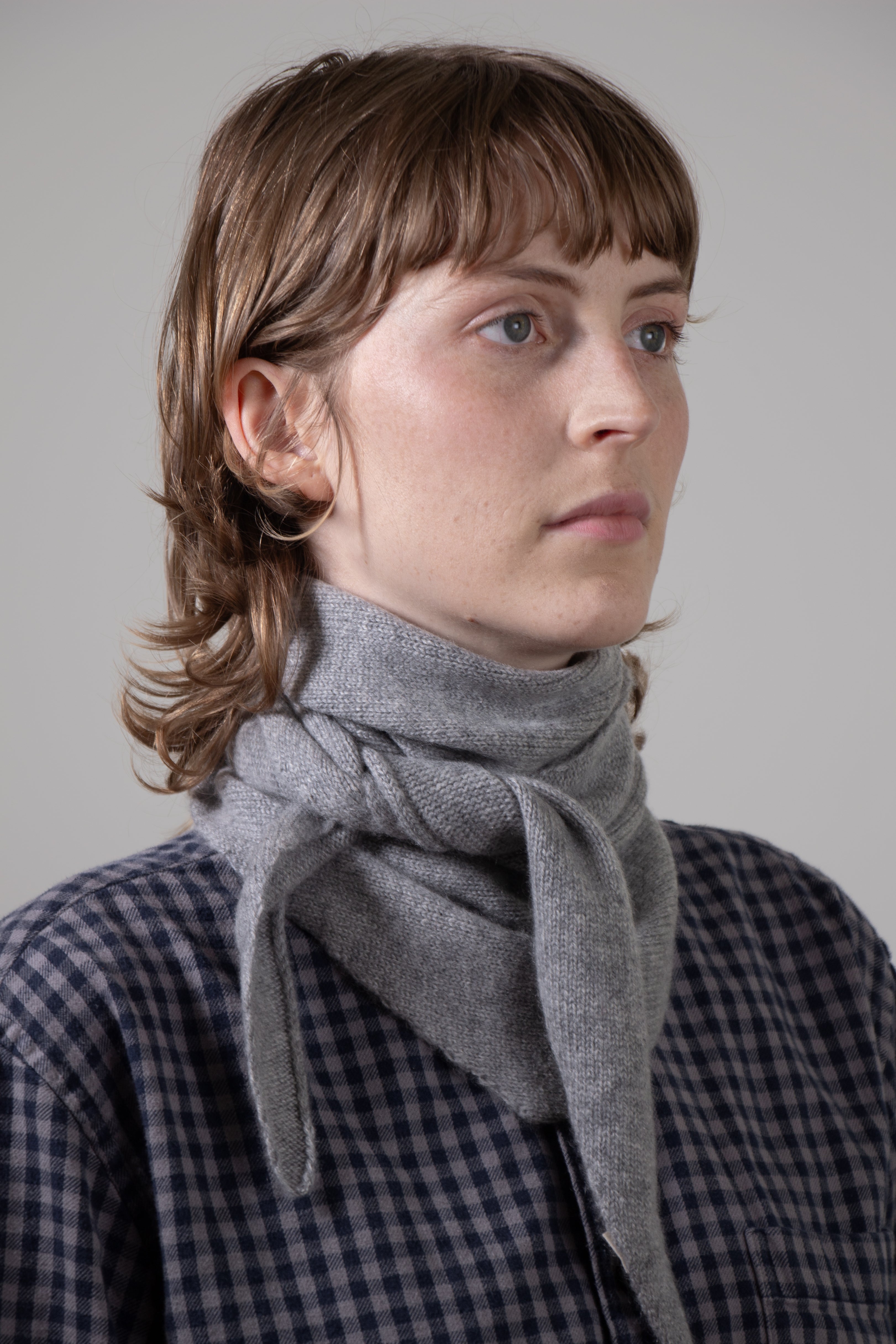 Person wearing a gray triangle scarf and checkered shirt against a plain background